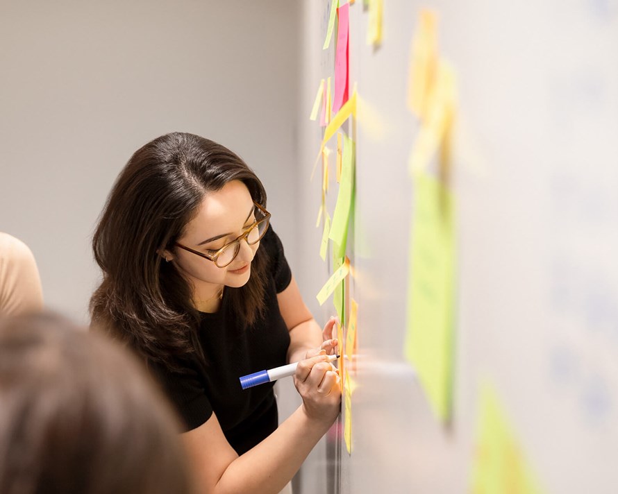 Woman with dark hair writing on a white board with post-it notes while two others collaborate 